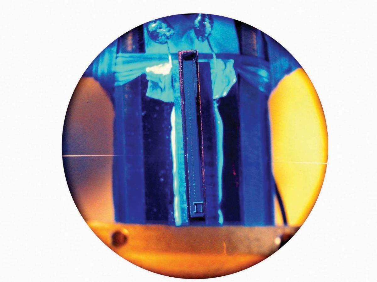 A quantum optic experimental system developed by Dr. Barak Dayan at Caltech. An optic fiber (horizontal line, left) brings light to resonators (light dots appearing in a vertical row on the optic chip, center). Lasers above the chip are used to cool and manipulate single atoms